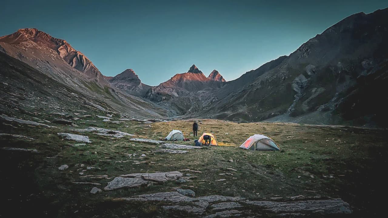 Alpine bivouac under the Aiguilles d'Arves, tents lit up at dusk, majestic panorama.