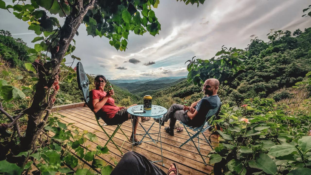 Two people relax on a wooden terrace, surrounded by greenery, admiring a mountainous landscape.