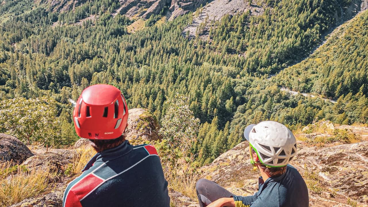 Two people in helmets admire a spectacular view of a wooded valley from a cliff.