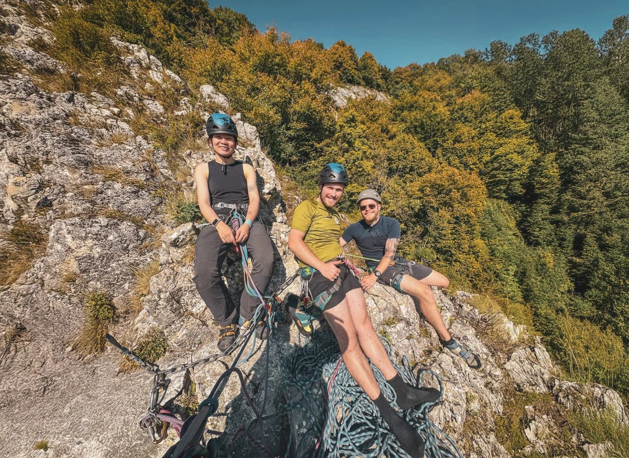 Three smiling climbers rest on a sunny cliff, surrounded by lush nature.