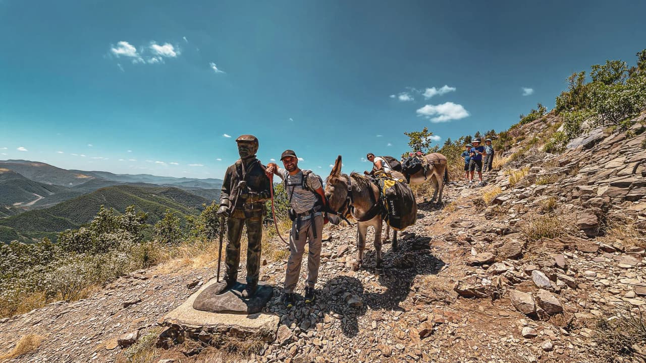 Hikers accompanied by donkeys on a sunny path, statue of Stevenson, panoramic view.