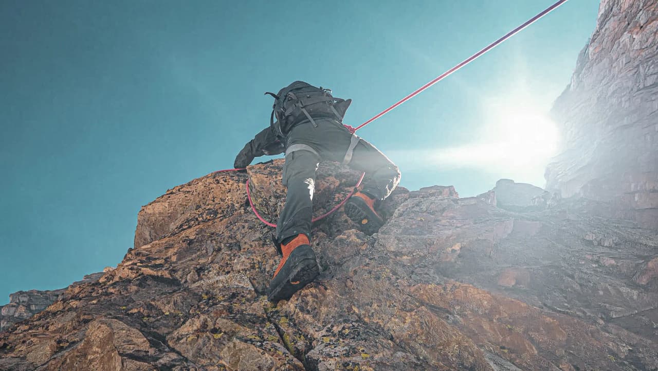Climber climbing a rock under a blue sky, bright light in the background.