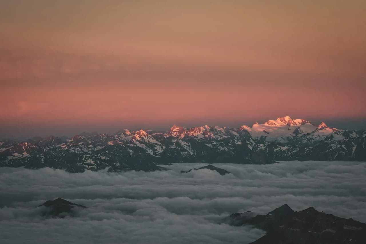 Alpine landscape at sunset, snow-capped mountains emerging from the clouds.