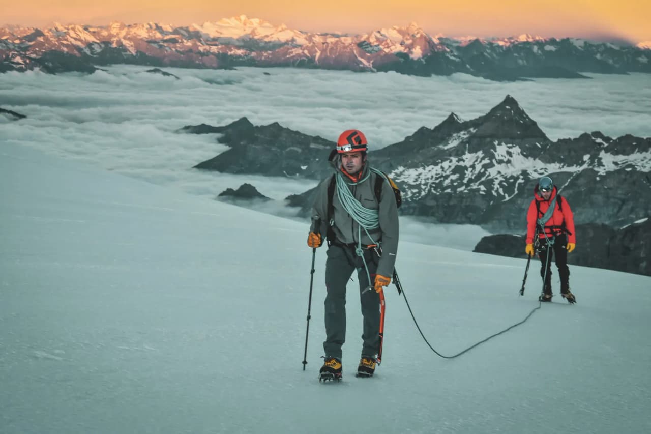 Two climbers make their way across a glacier, with a majestic mountain landscape in the background.