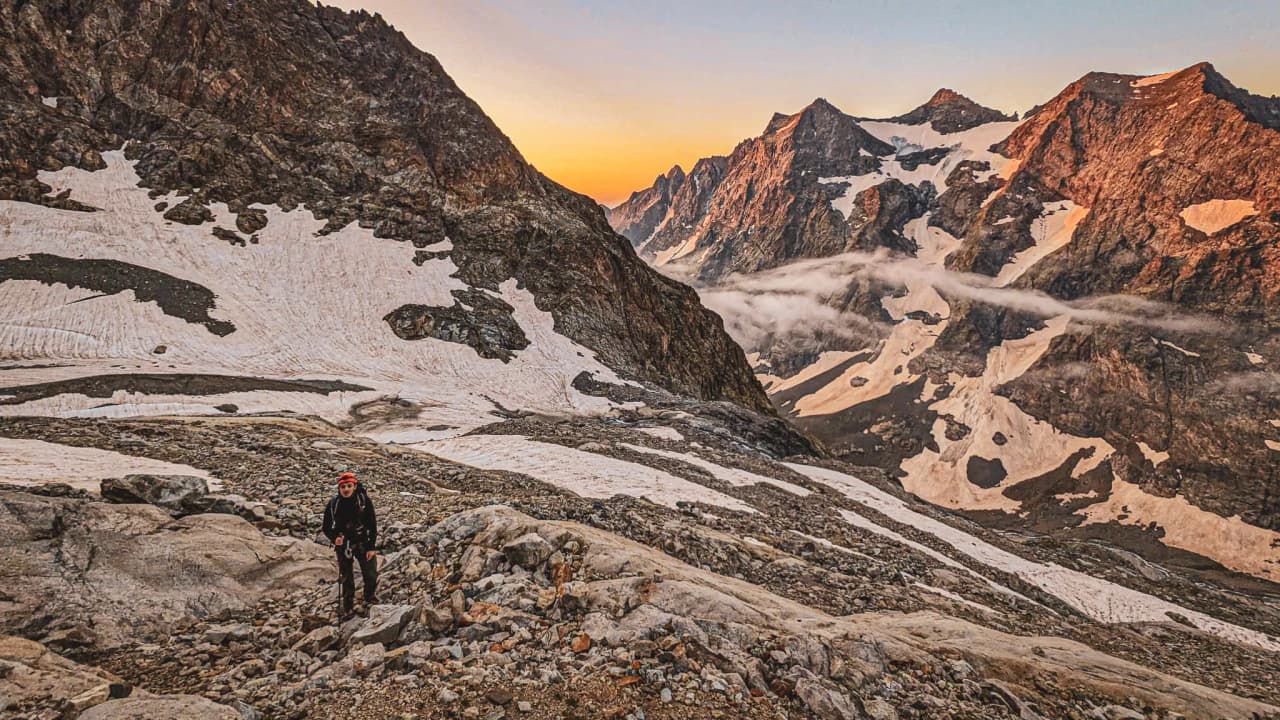 A mountaineer climbs a rocky path, surrounded by the majestic peaks of the Écrins at sunrise.