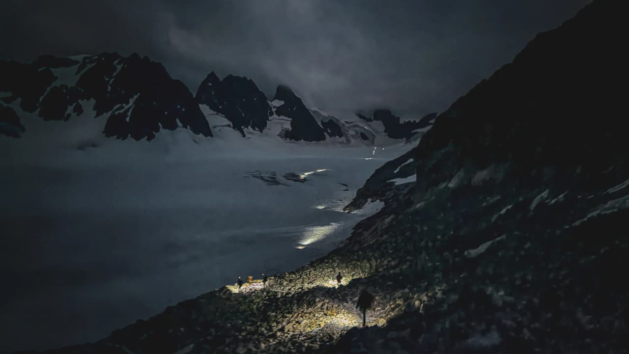 A night-time expedition in the mountains, with climbers illuminated by headlamps on a rocky path, surrounded by majestic snow-capped peaks.