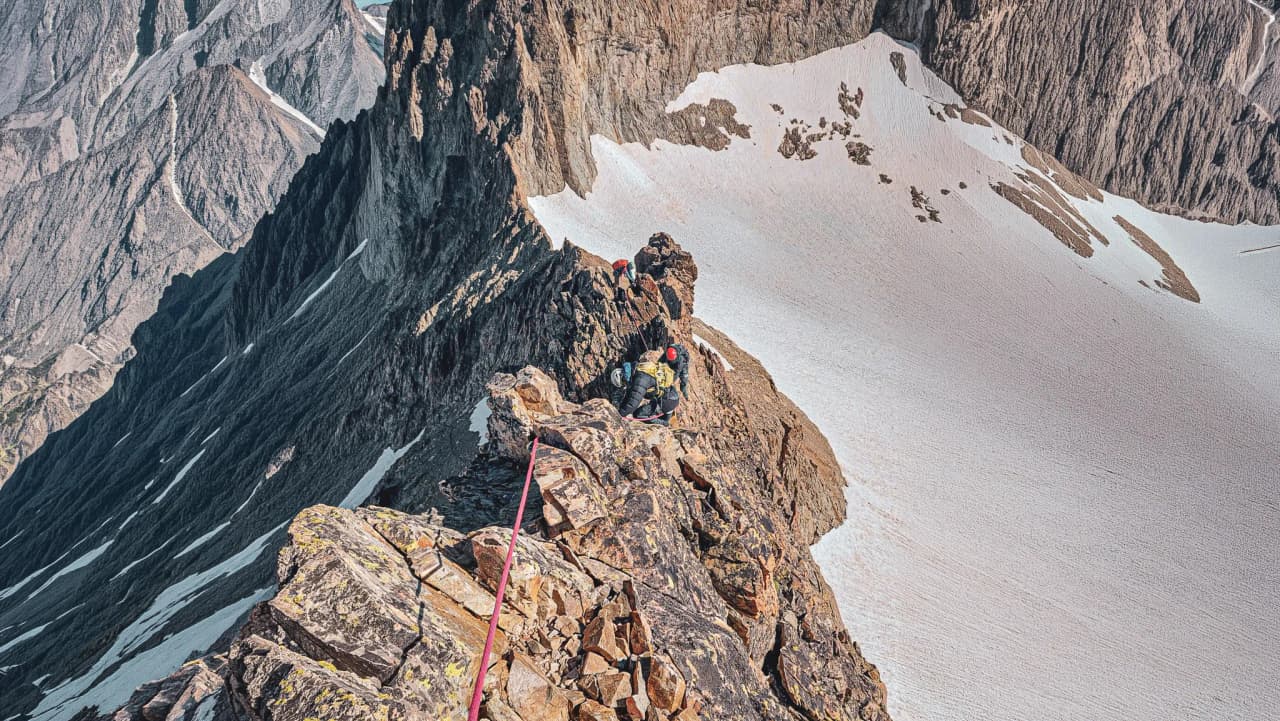 Climbers on a steep ridge, with majestic peaks in the background and sparkling snow.