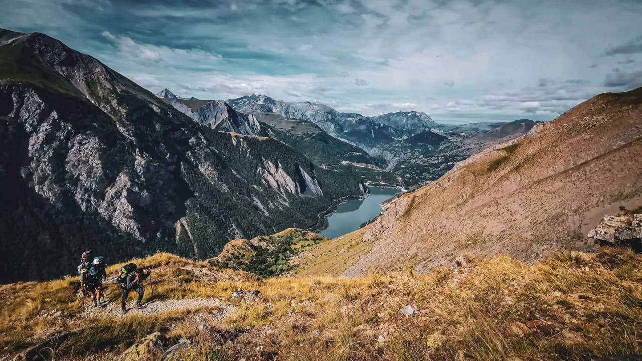 A breathtaking view of the mountains with hikers, greenery and a lake glistening under the blue sky.