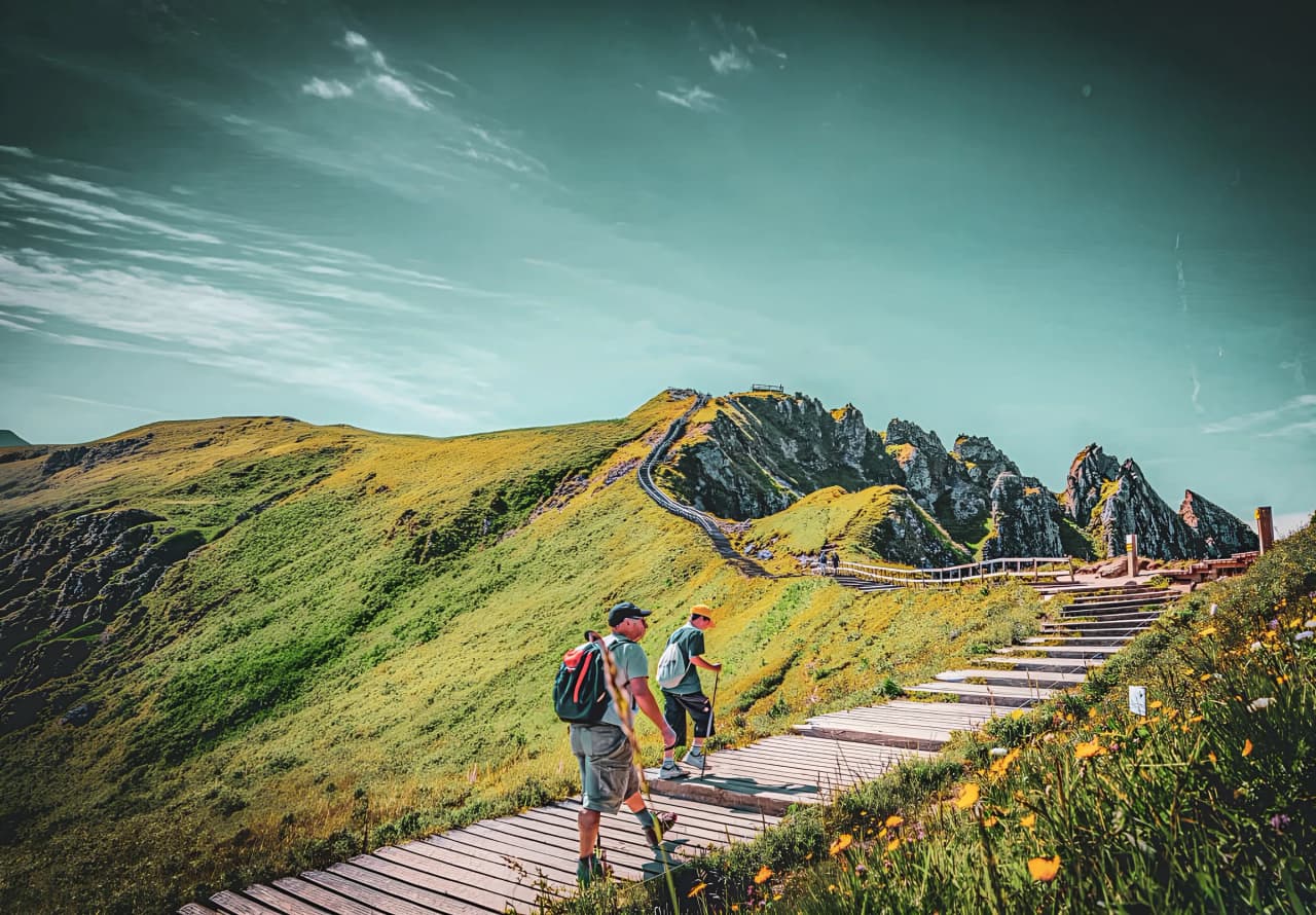 Two hikers on a path amidst the lush green ridges of the Massif Central, clear skies.