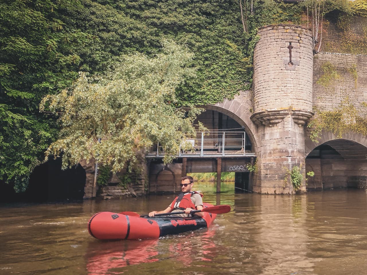 Un pagayeur en packraft navigue paisiblement sous un ancien pont verdoyant en Belgique.