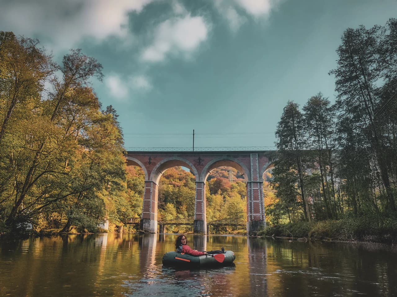 Expédition en packraft sur une rivière entourée de paysages d'automne et d'un viaduc majestueux.