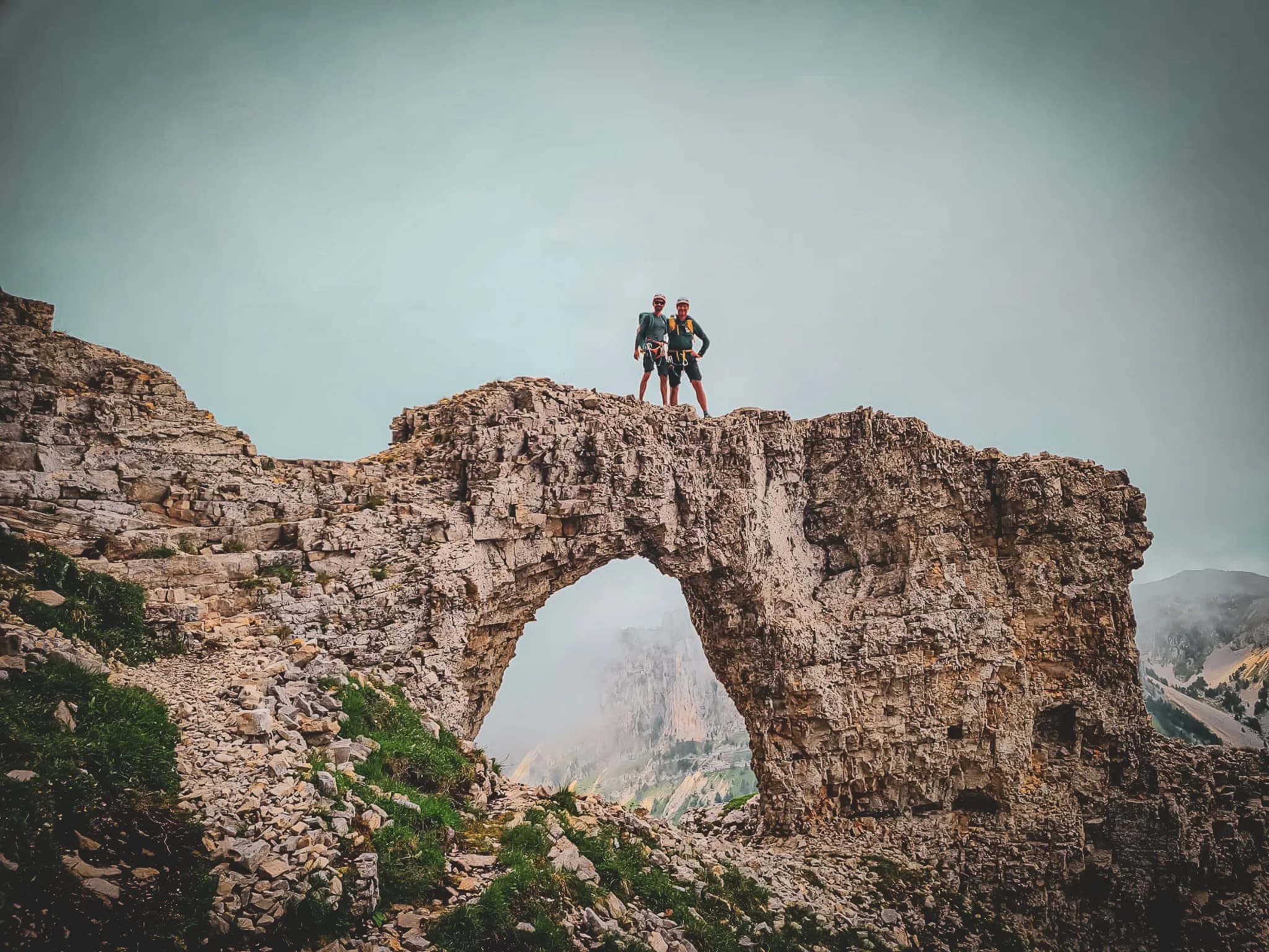 Two hikers pose on a rocky arch, with a breathtaking panorama of the mountains.