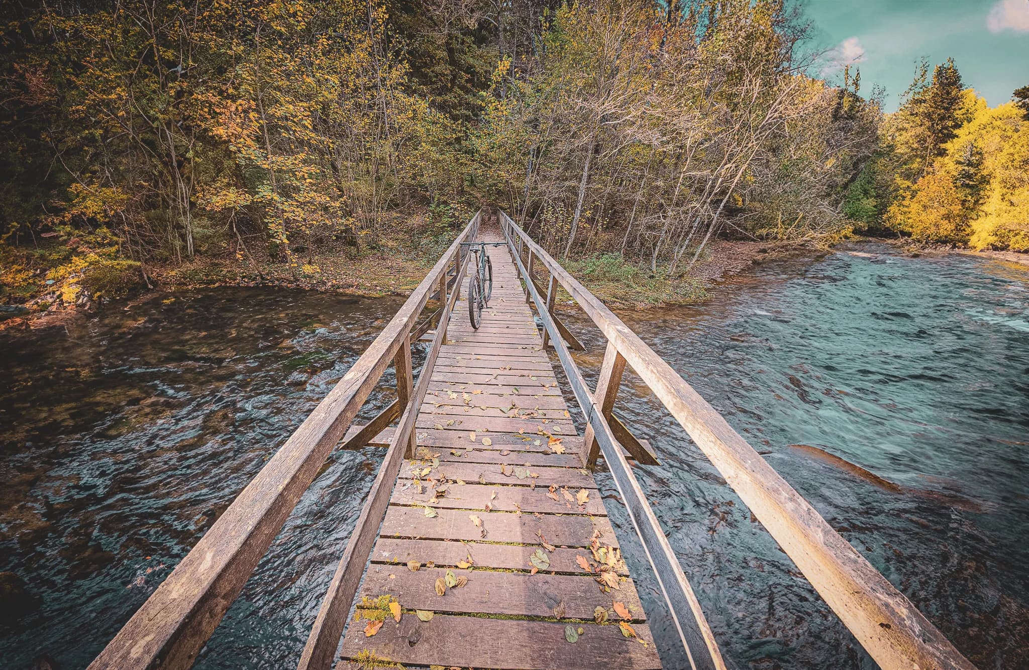 A wooden bridge crossing a river, lined with leaves and surrounding vegetation. Trees with colourful foliage, typical of autumn, frame the path. A bicycle is placed on the bridge.