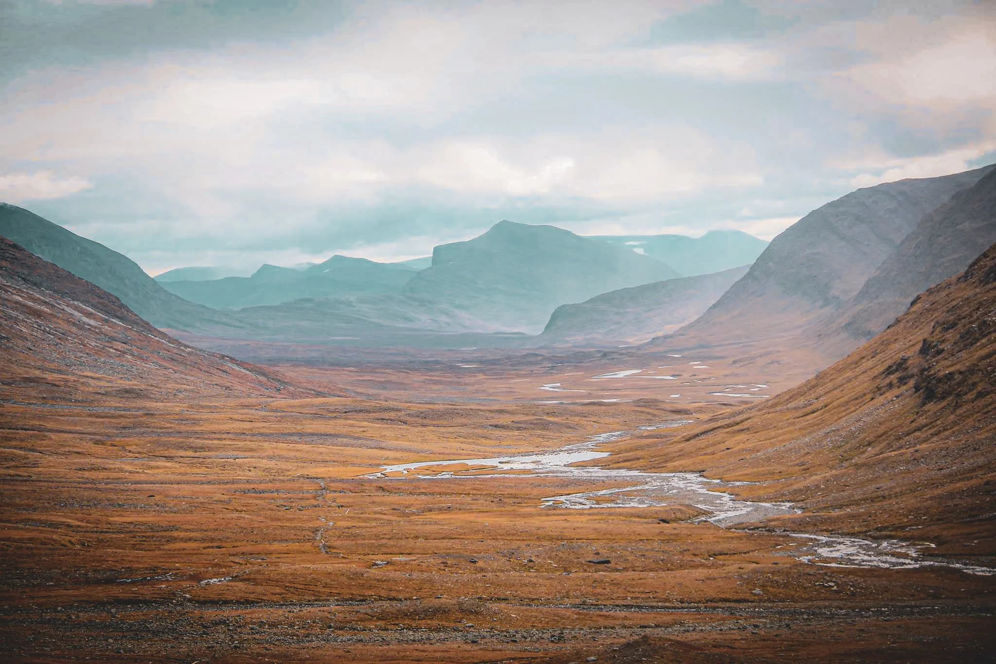 Un paysage vaste et polaire, avec des vallées étendues recouvertes d'herbe dorée et des montagnes en arrière-plan. Un ruisseau sinueux traverse le terrain, ajoutant une touche de luminosité.