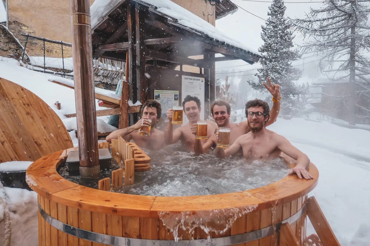 Four friends relax in an outdoor hot tub, surrounded by snow and mountains.