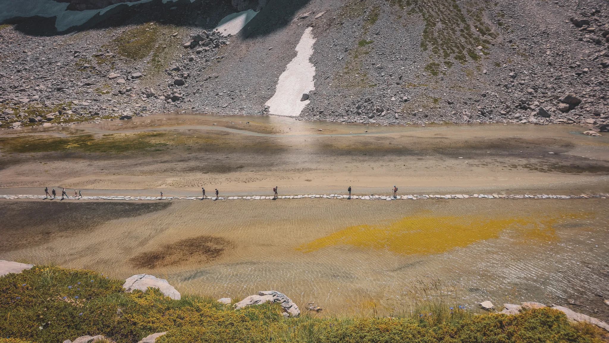 A natural landscape. In the centre, a stone path stretches across a shallow stream, where people are walking. The surface of the water is clear, allowing the background to be seen, while shades of yellow and