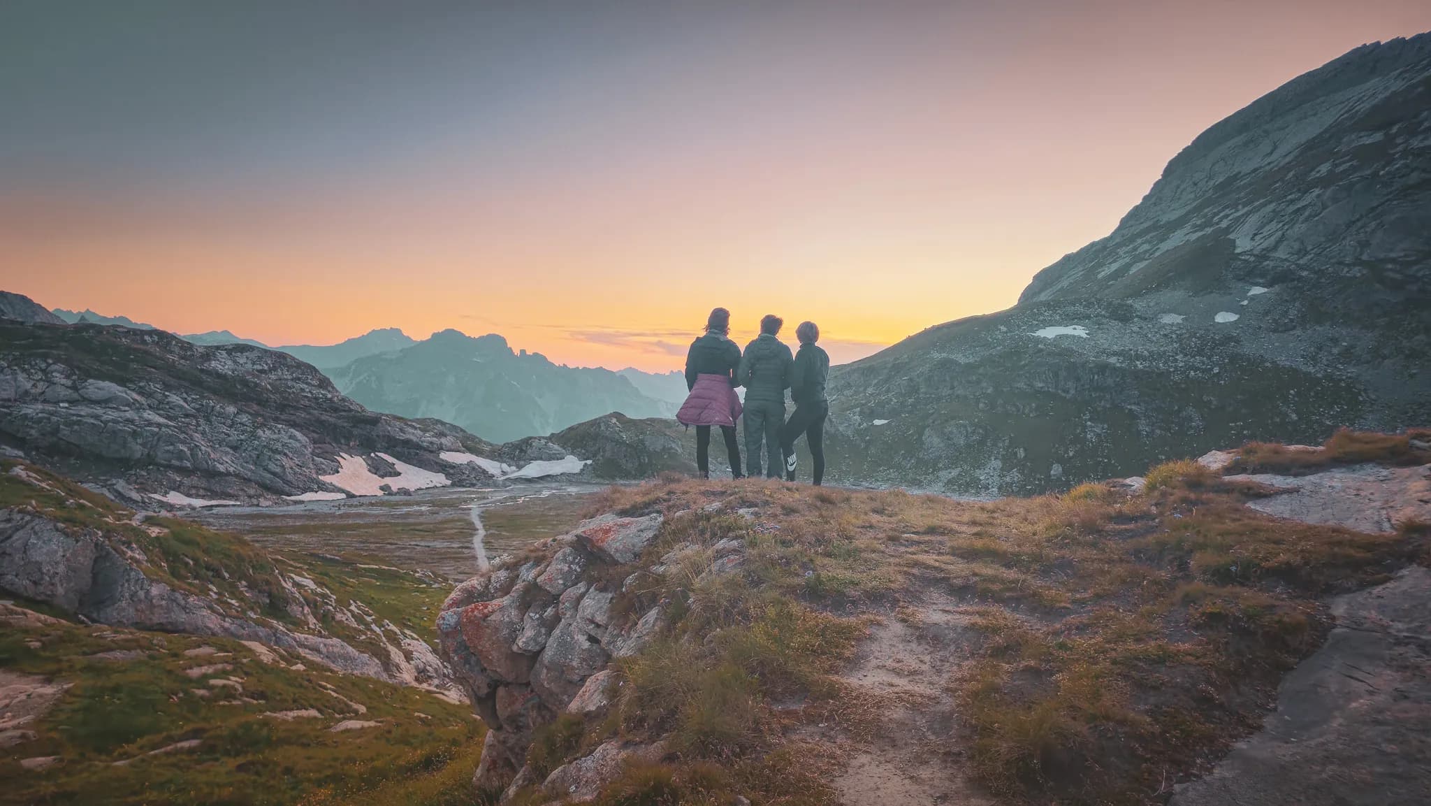 A group of three people stand on the edge of a cliff, admiring a mountainous landscape at sunset. The silhouettes of the mountains can be seen in the background, bathed in warm shades of pink and orange.