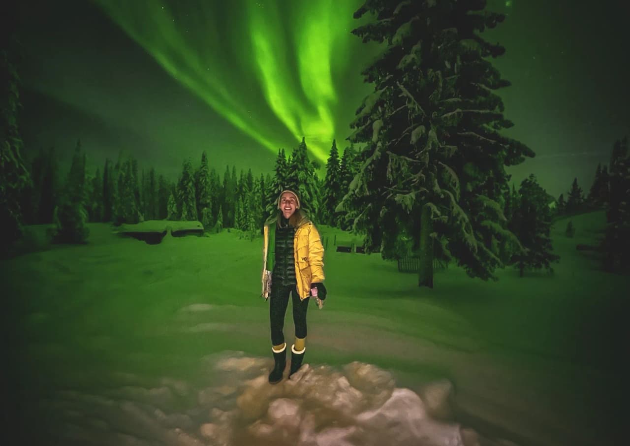 A smiling woman under the northern lights, surrounded by snow and fir trees in Lapland.