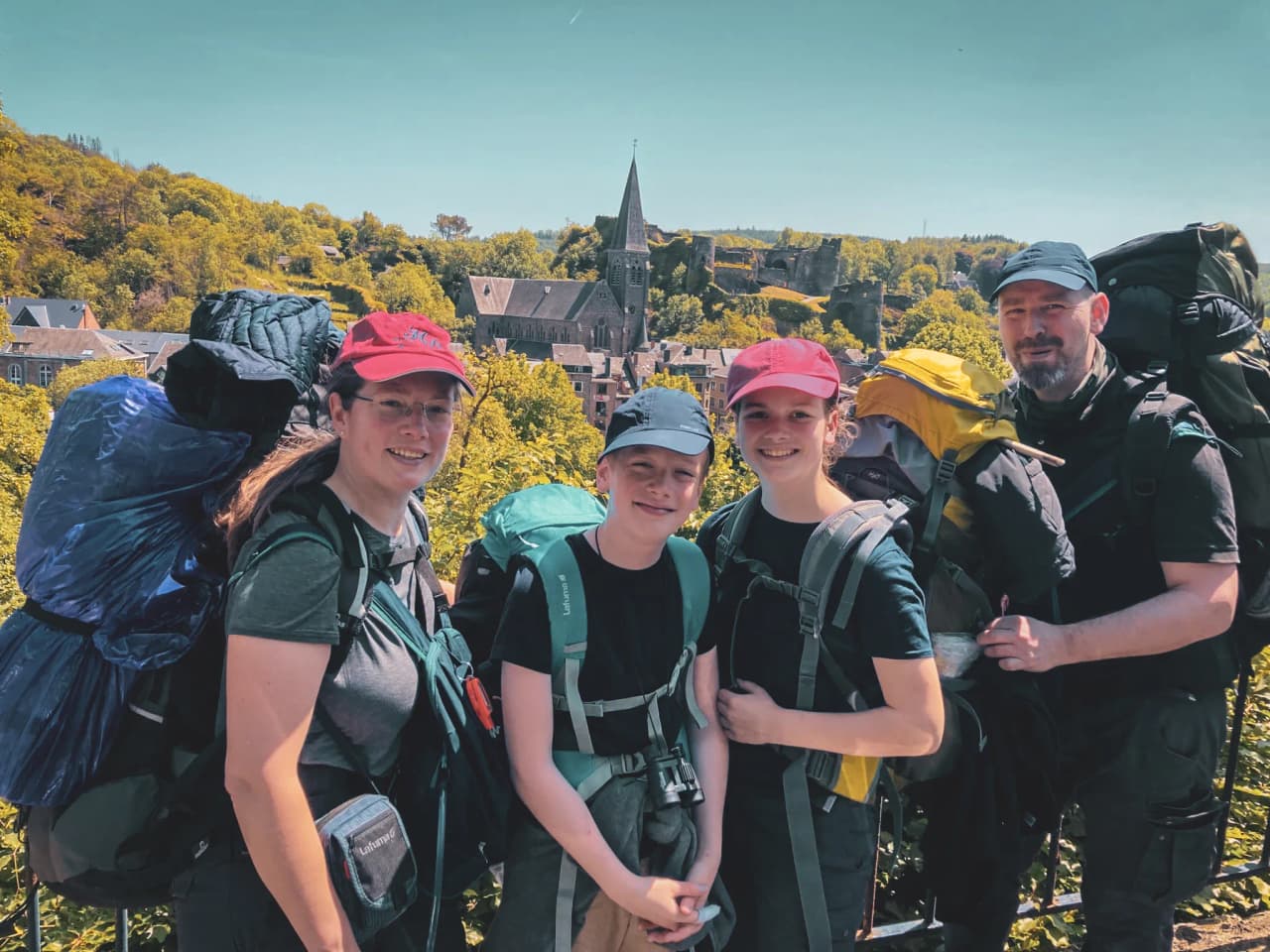 Une famille souriante, équipée de sacs à dos, devant un village pittoresque en pleine nature.