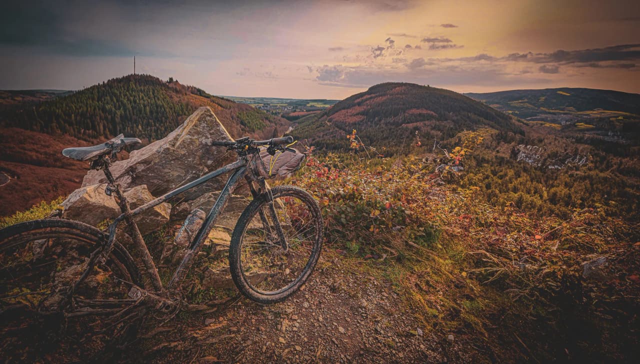 A mountain bike is leaning against a rock at the edge of a path overlooking a hilly landscape. The scene is bathed in the golden light of a late day, with hills blanketed by the sun.