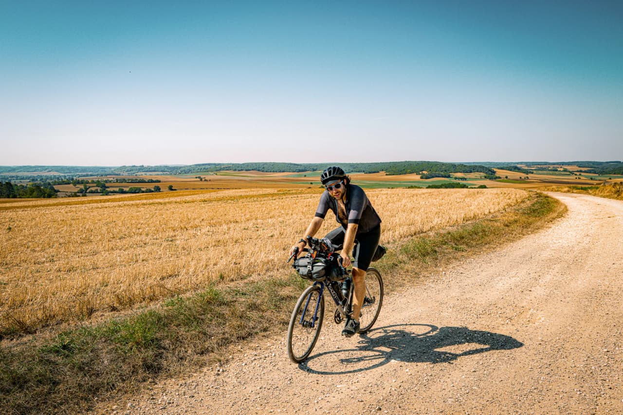 Cyclist on a dirt road through golden fields, under a sunny blue sky.