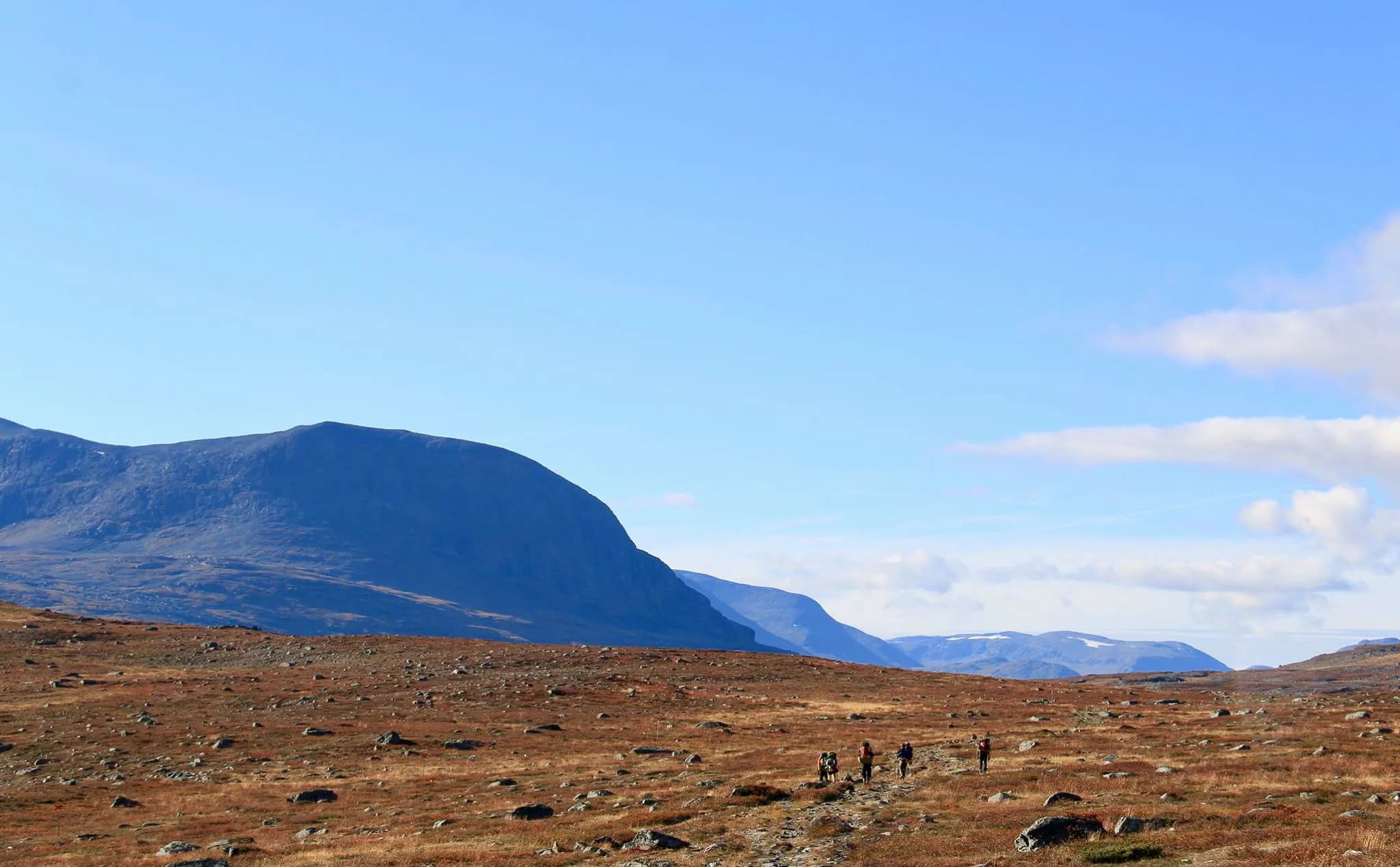 Un paysage montagneux avec des collines et des pics en arrière-plan, sous un ciel bleu clair parsemé de quelques nuages blancs. Au premier plan, une vaste étendue de terrain aride et légèrement vallonné.