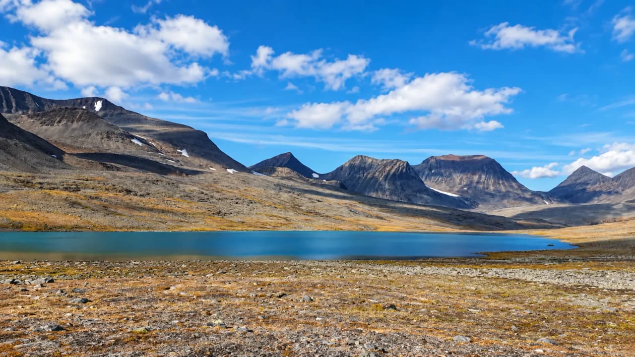 Un paysage montagneux majestueux en Laponie, avec un lac turquoise sous un ciel bleu parsemé de nuages.