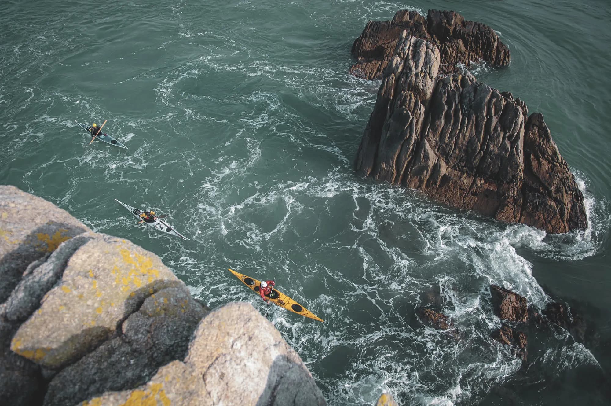 Three kayaks glide over rough waters, surrounded by rocks in the Molène archipelago.