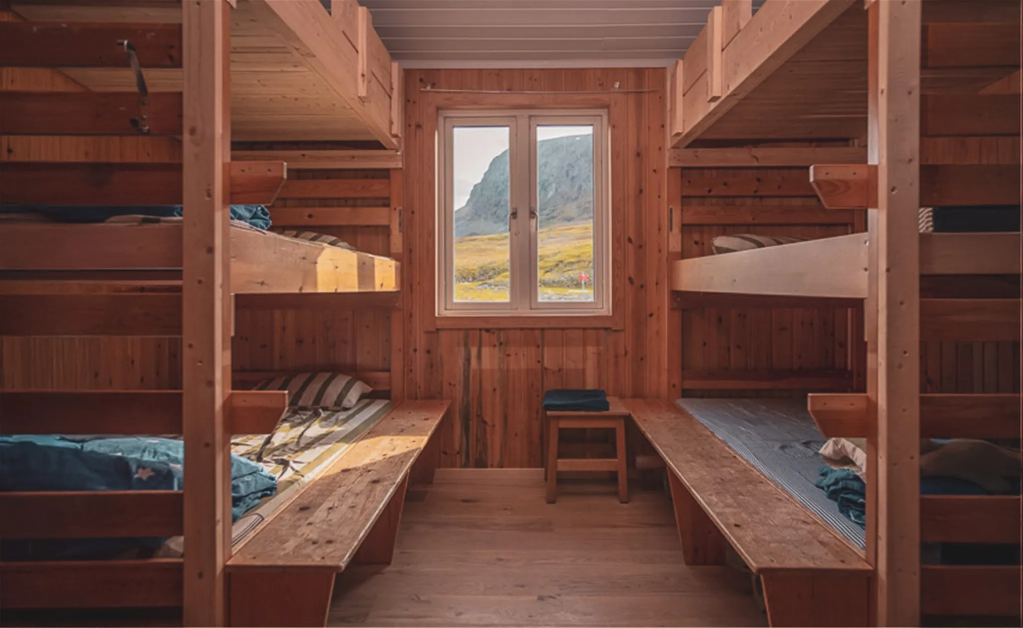 Une chambre en bois avec des lits superposés, offrant une ambiance chaleureuse et rustique. Les murs et le mobilier sont en bois, créant un espace naturel. Une fenêtre au centre donne sur un paysage montagneux, illuminant la pièce.