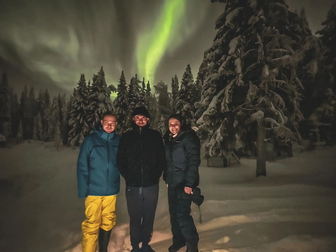 Three smiling people under the northern lights, surrounded by snowy landscapes in Lapland.