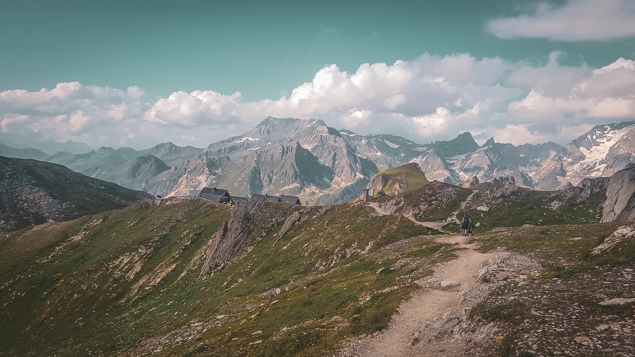 A captivating mountain landscape, characterised by majestic peaks shrouded in light clouds. A winding path cuts through lush greenery, while steep ridges rise up in the background.