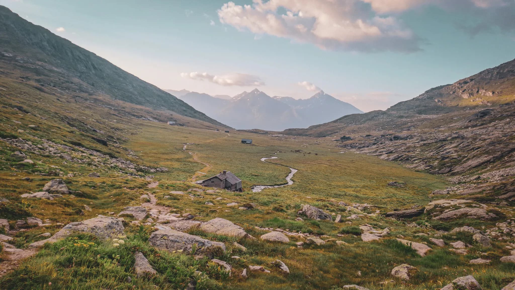 A wooden chalet stands at the foot of a majestic mountain, surrounded by greenery. The mountain, with its steep slopes and snow-covered areas, dominates the landscape, creating a striking contrast with the surrounding countryside.