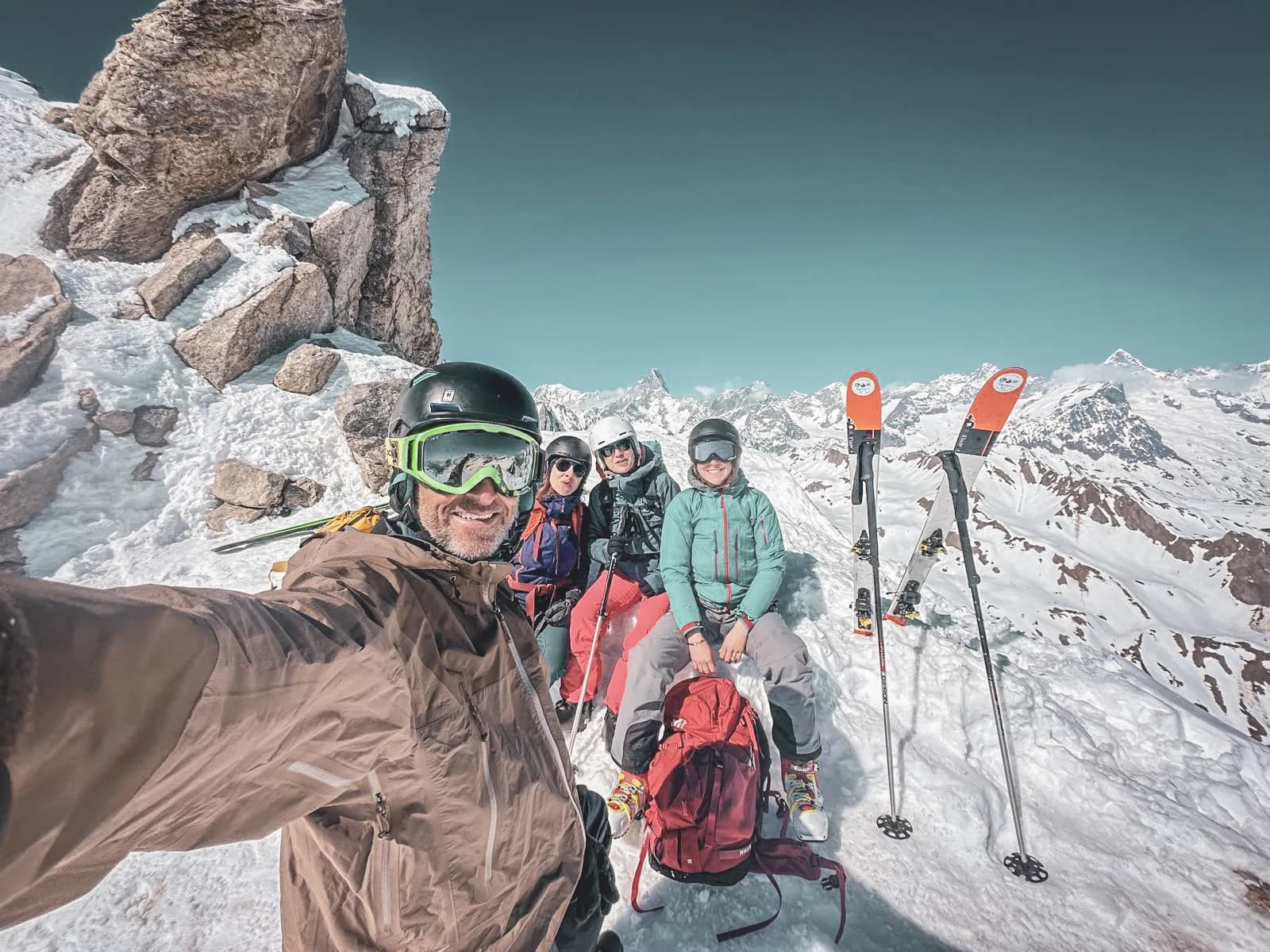 Four friends soak up the sun on an Alpine summit, skis in hand, with majestic scenery in the background.