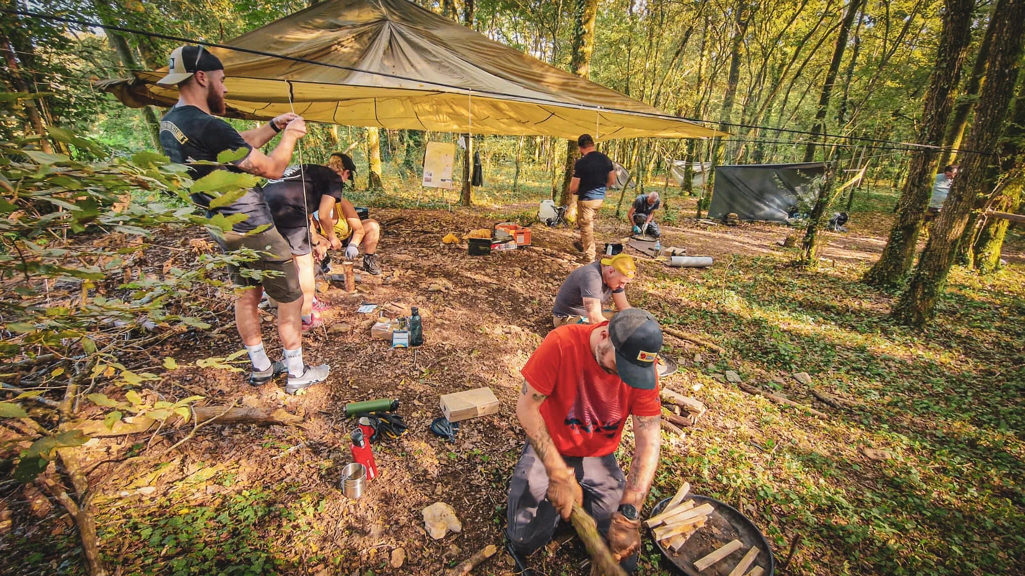 Groupe en pleine session de survie en forêt, créant abris et feu sous un ciel ensoleillé.