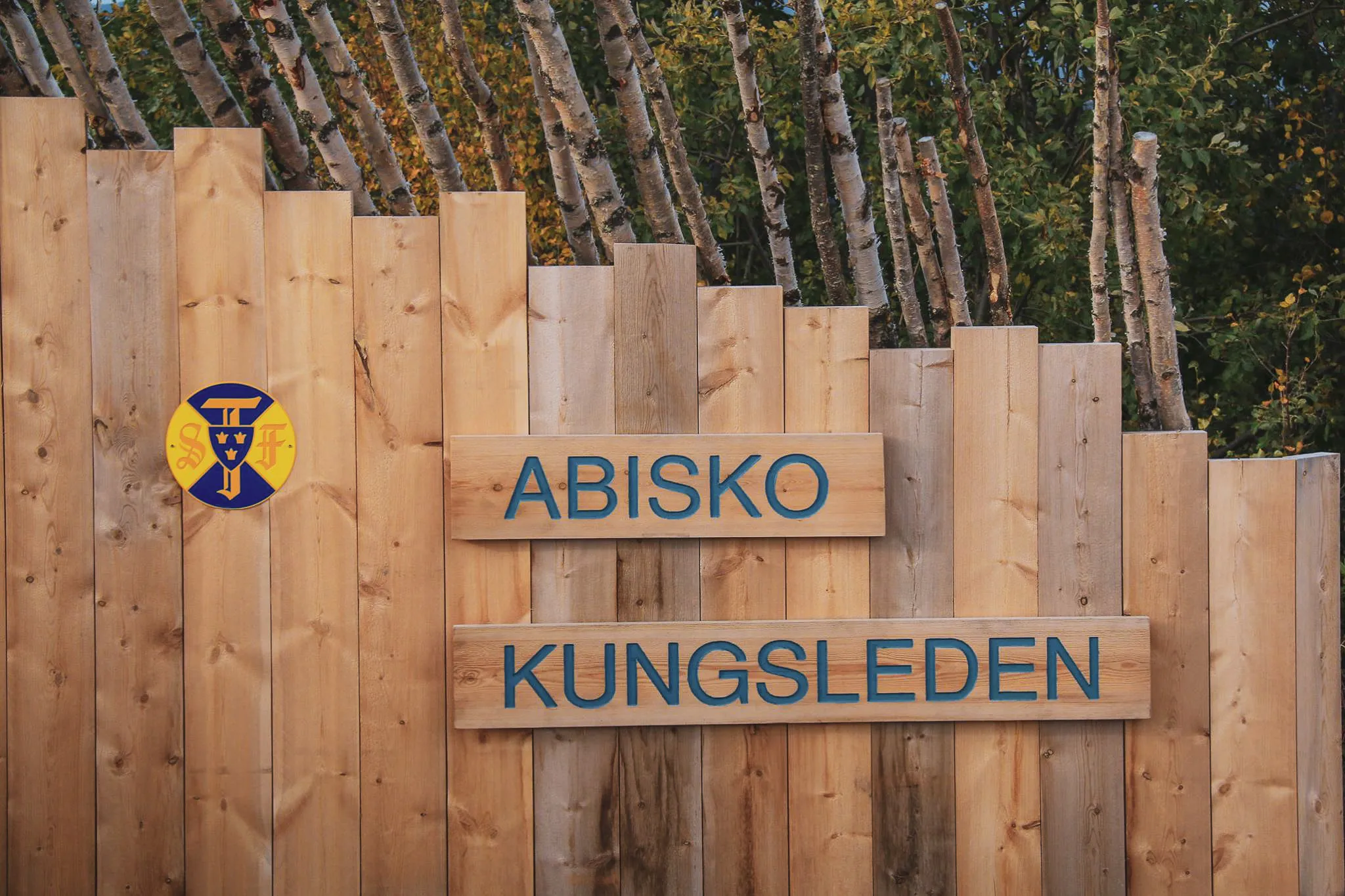 Un panneau en bois stylisé présente les inscriptions "ABISKO" et "KUNGSLEDEN" en lettres bleues, entouré par des planches de bois aux teintes variées.
