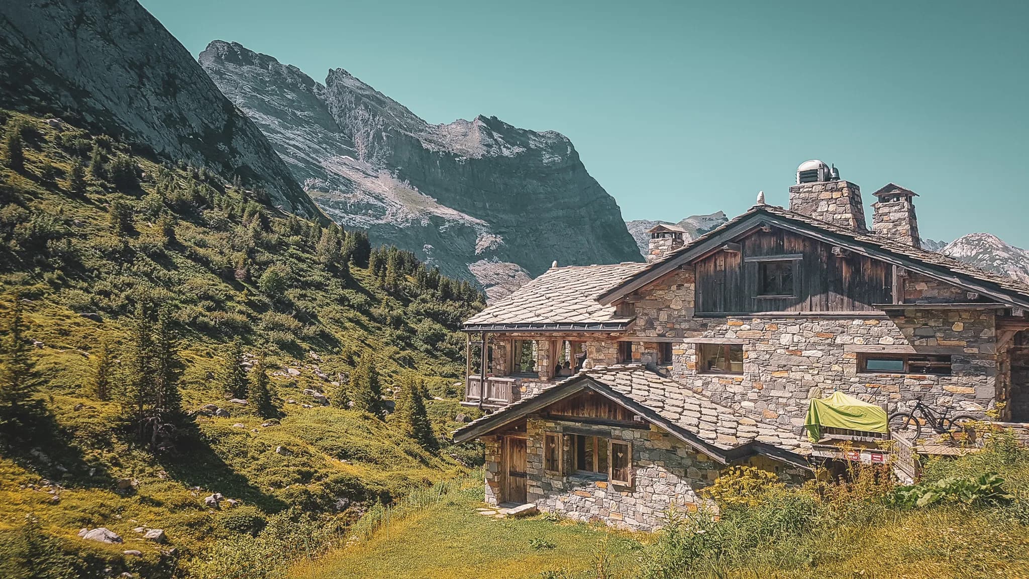 An elegant stone chalet nestled at the foot of majestic mountains. The building features a slate roof and wooden beams, creating a rustic charm. The surroundings are lush with trees and plants.