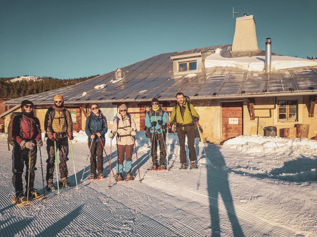 A group of adventurers on snowshoes in front of a snow-covered Mountain hut in the Swiss Jura, under a clear sky.