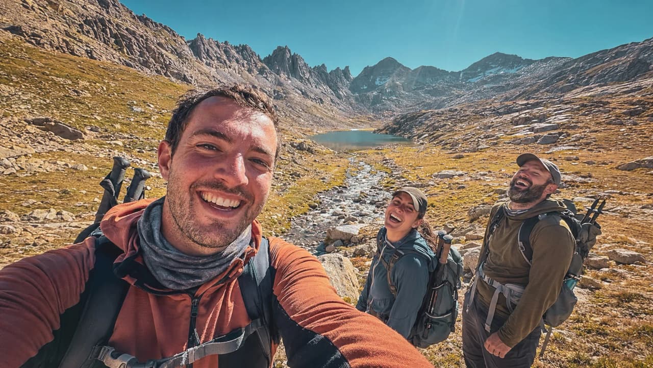 Trois randonneurs souriants devant un lac alpin et des montagnes majestueuses.