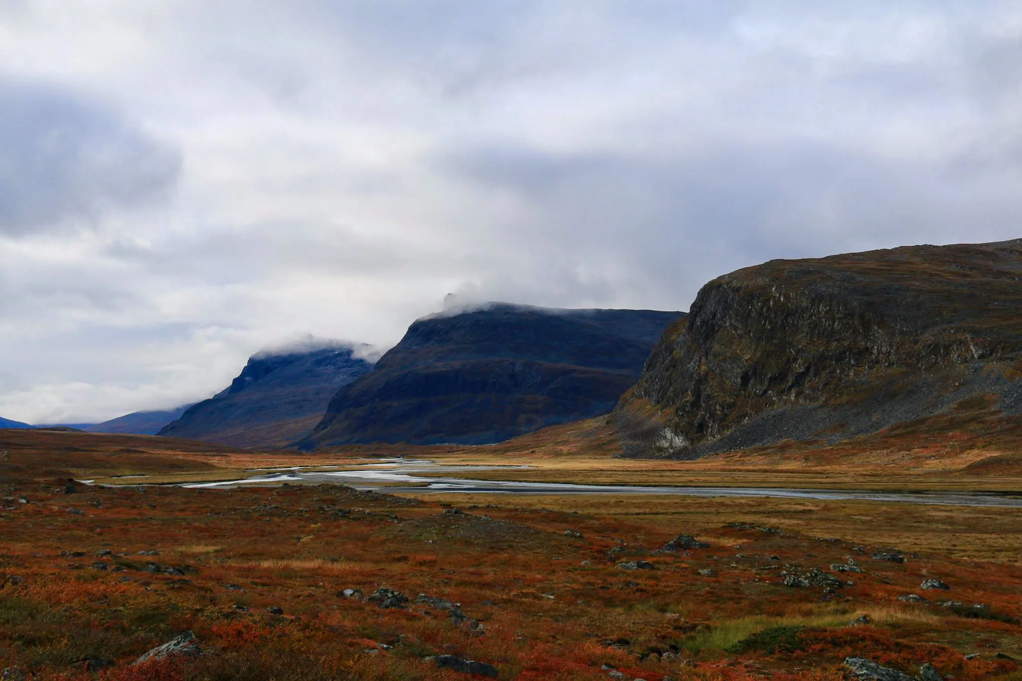 Un paysage montagneux avec des collines escarpées, partiellement couvertes de nuages. Au premier plan, une vallée verdoyante parsemée de rochers et de buissons, arborant des teintes rouges et orangées.