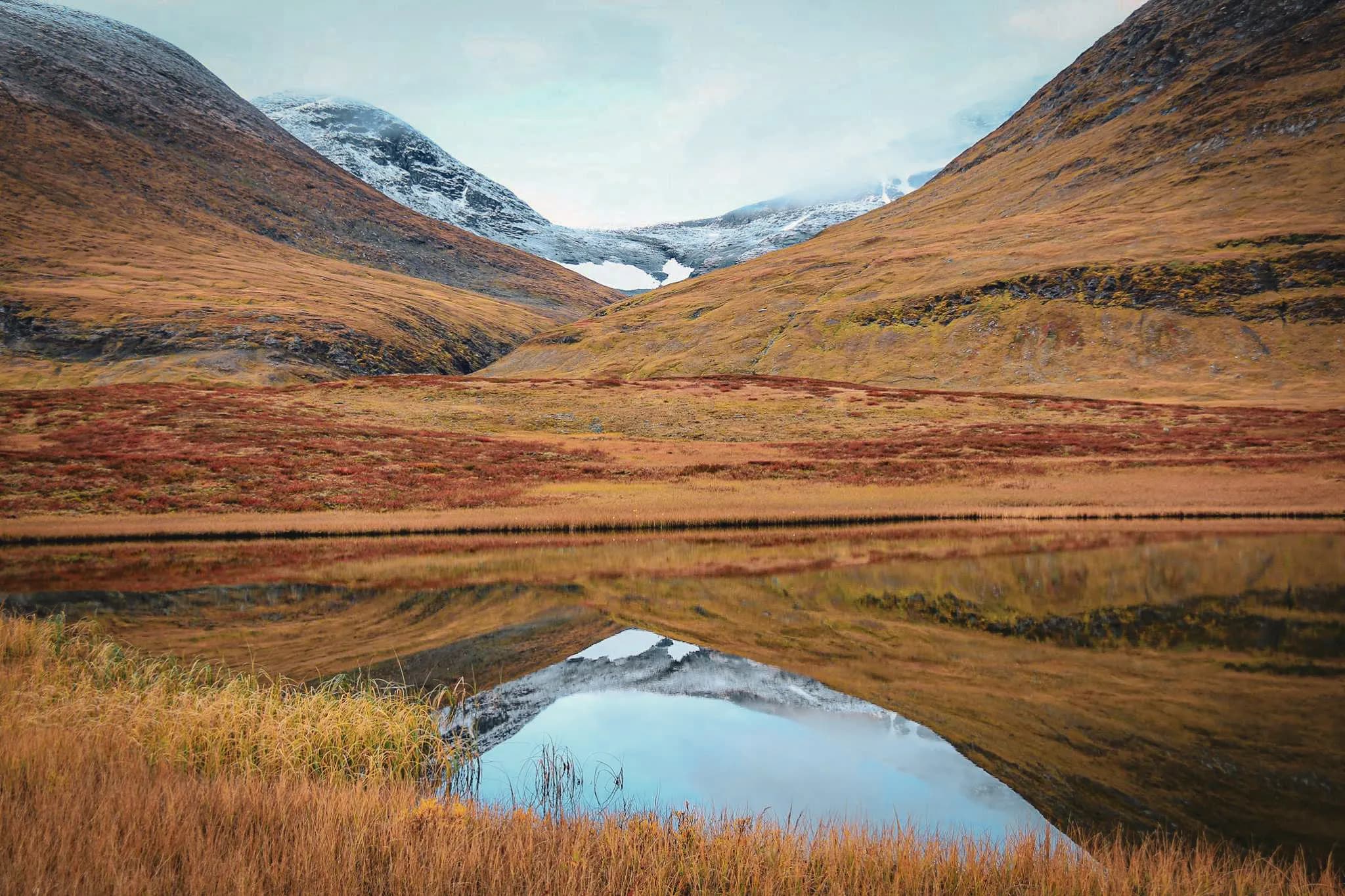 Un paysage montagnard présentant des collines aux teintes dorées et brunes, avec des sommets recouverts de neige au loin. À l'avant, un lac calme reflète les montagnes et le ciel nuageux.