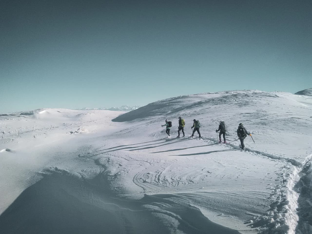 A group on snowshoes trek across a vast snow-covered landscape in the Swiss Jura under a blue sky.