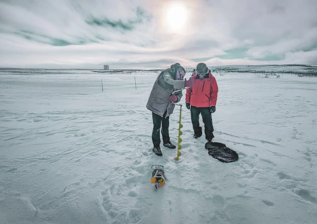 Two people drill through the ice in a picturesque snowy landscape, exploring Norwegian Lapland.