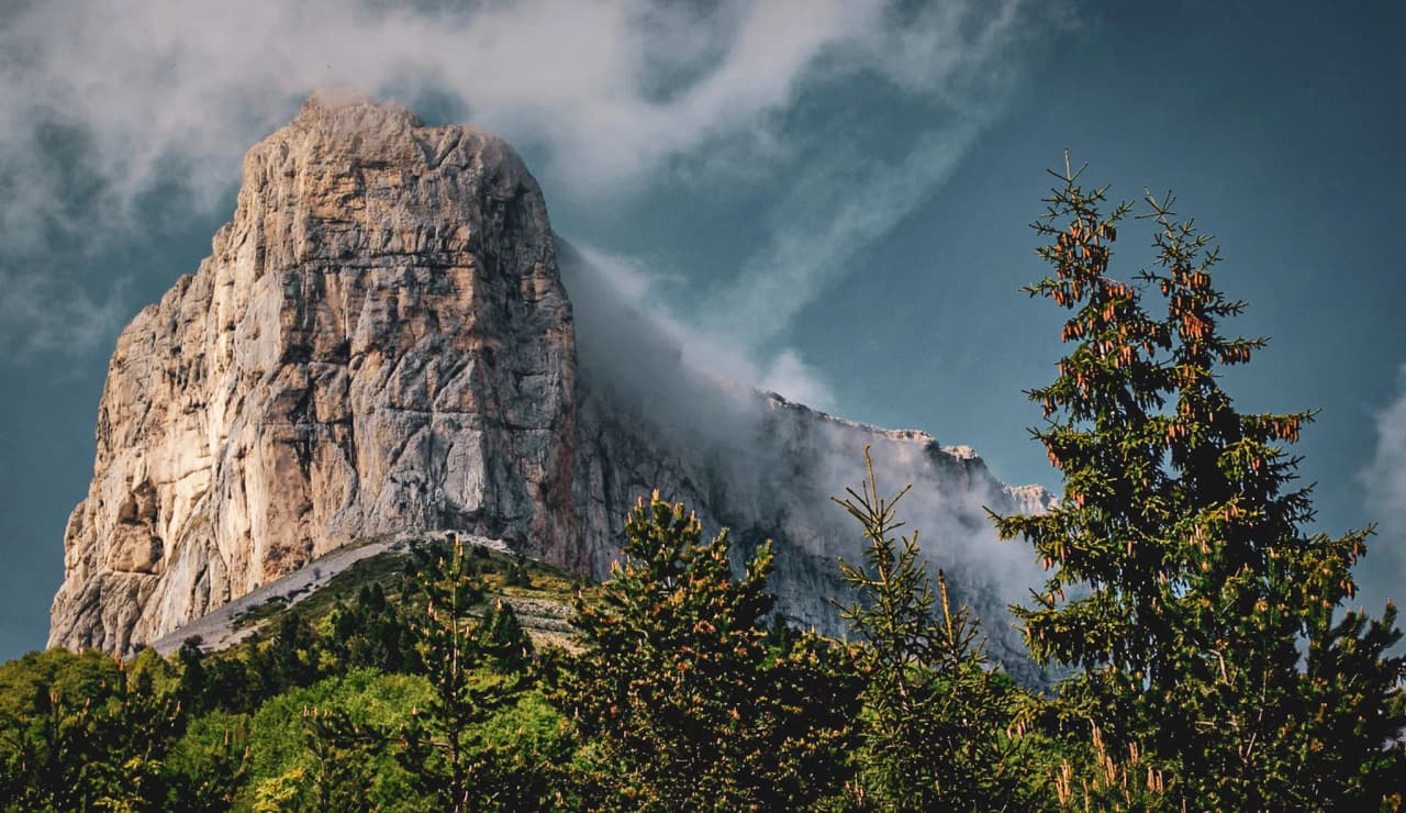 Majestic view of Mont Aiguille, surrounded by pine trees, under a blue sky dotted with clouds.