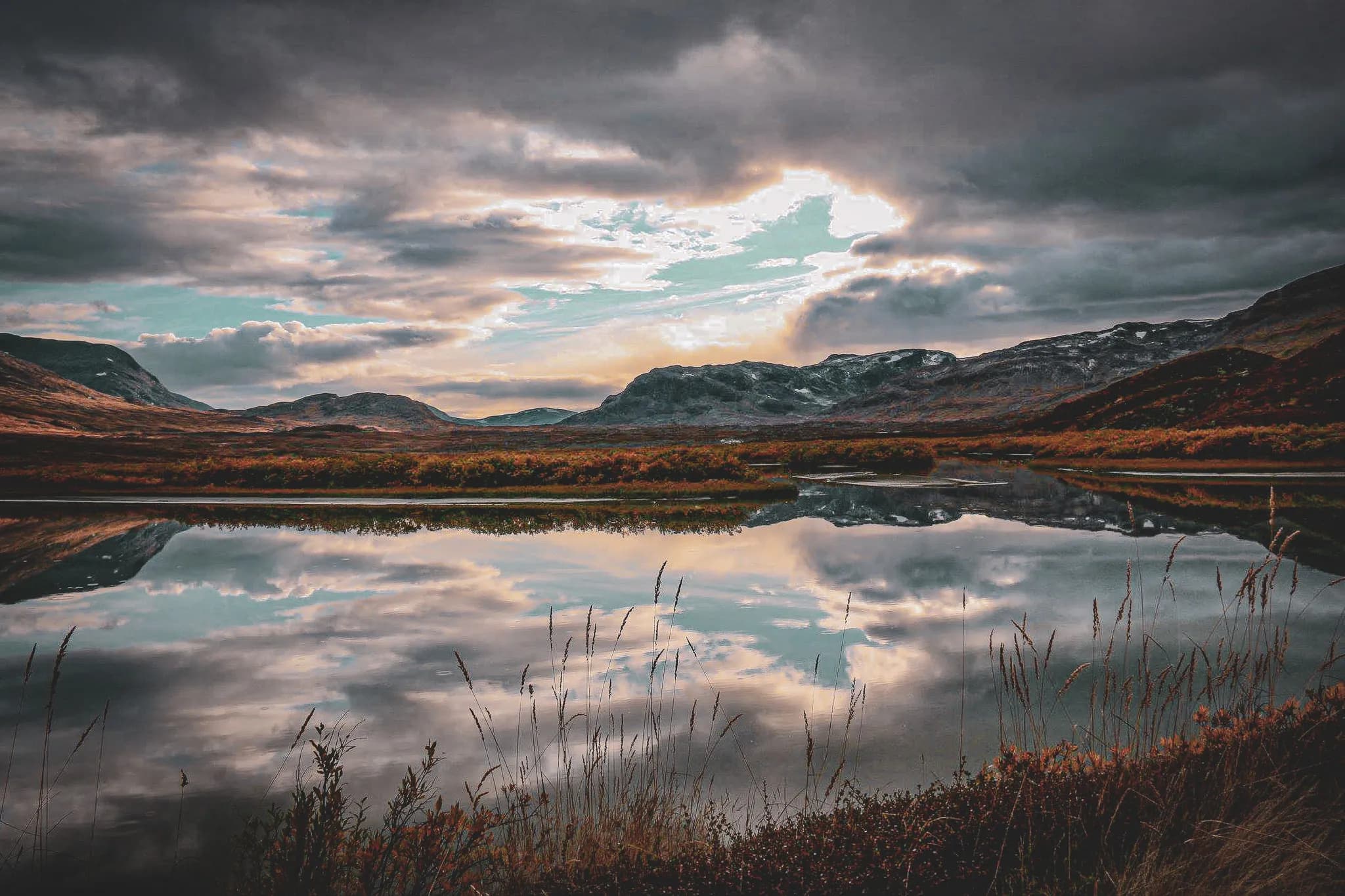 Un paysage montagneux au crépuscule, avec un lac calme réfléchissant les nuages et les collines environnantes. Les montagnes se dressent majestueusement à l'arrière-plan.