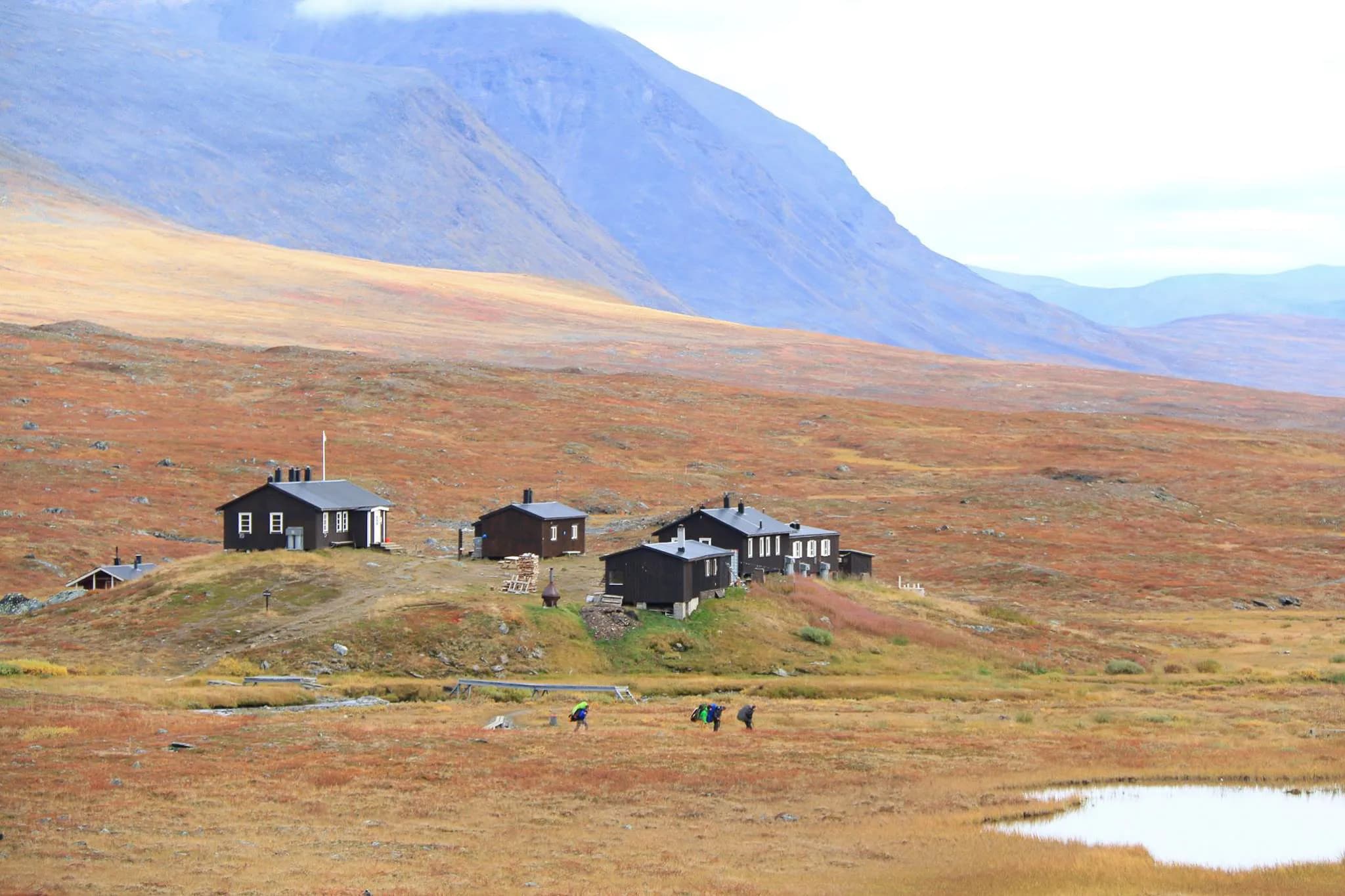 Un paysage de montagne avec des collines dorées et verdoyantes en arrière-plan, où se dressent plusieurs chalets en bois noir. Ces cabines STF sont réparties sur une légère élévation, entourées de prairies aux te