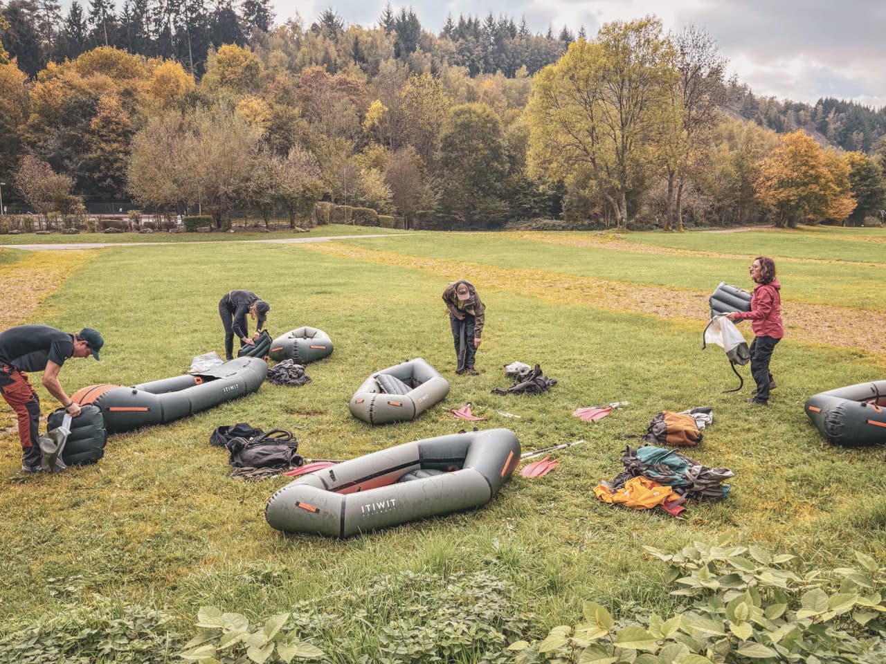 Un groupe prépare des packrafts sur un terrain verdoyant, entouré de forêts colorées en automne.