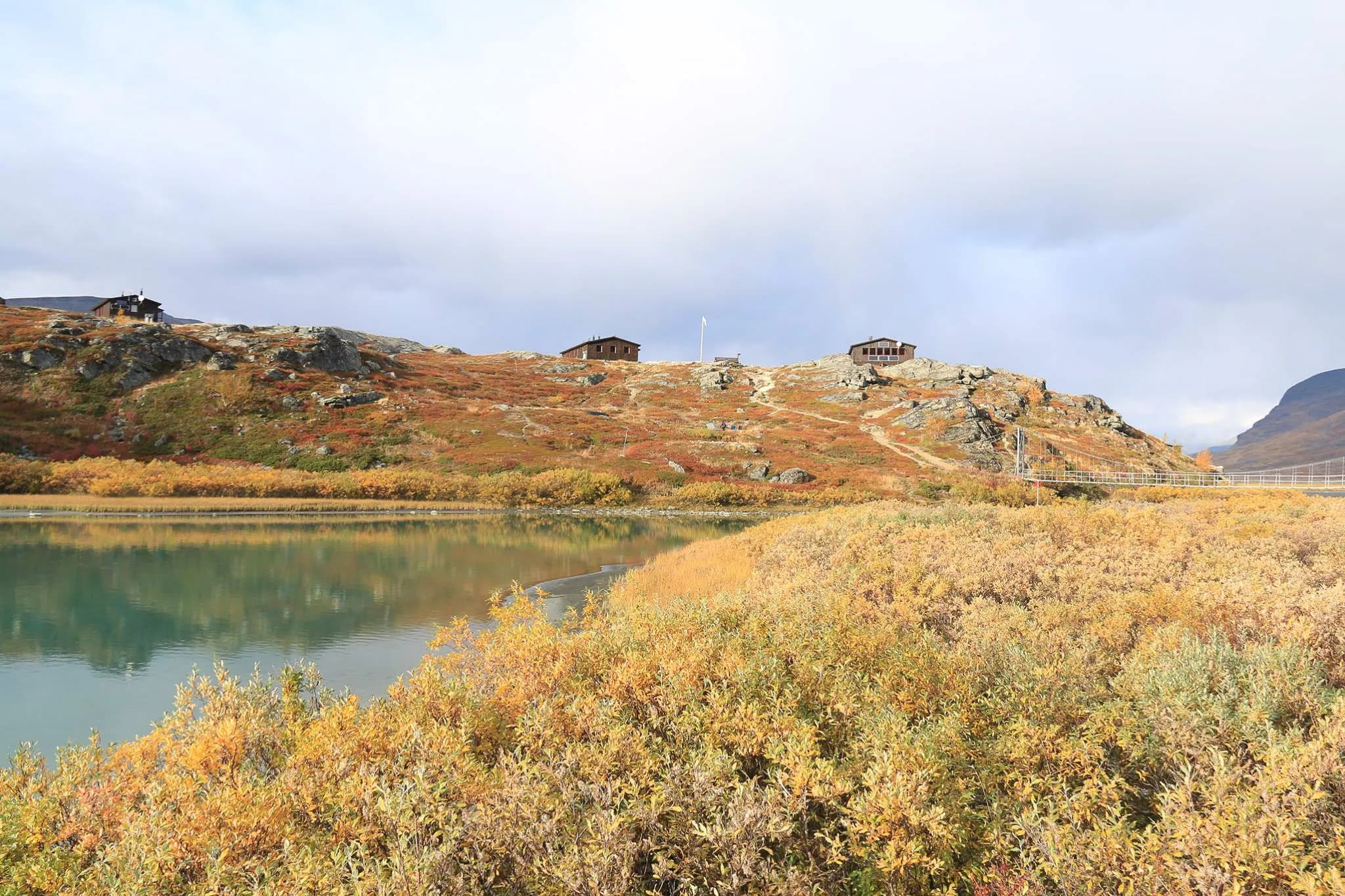 Un paysage Nordique paisible avec un lac au premier plan, entouré de feuillage automnal aux teintes dorées et orange. À l'arrière-plan, des chalets en bois sont perchés sur une colline rocailleuse.