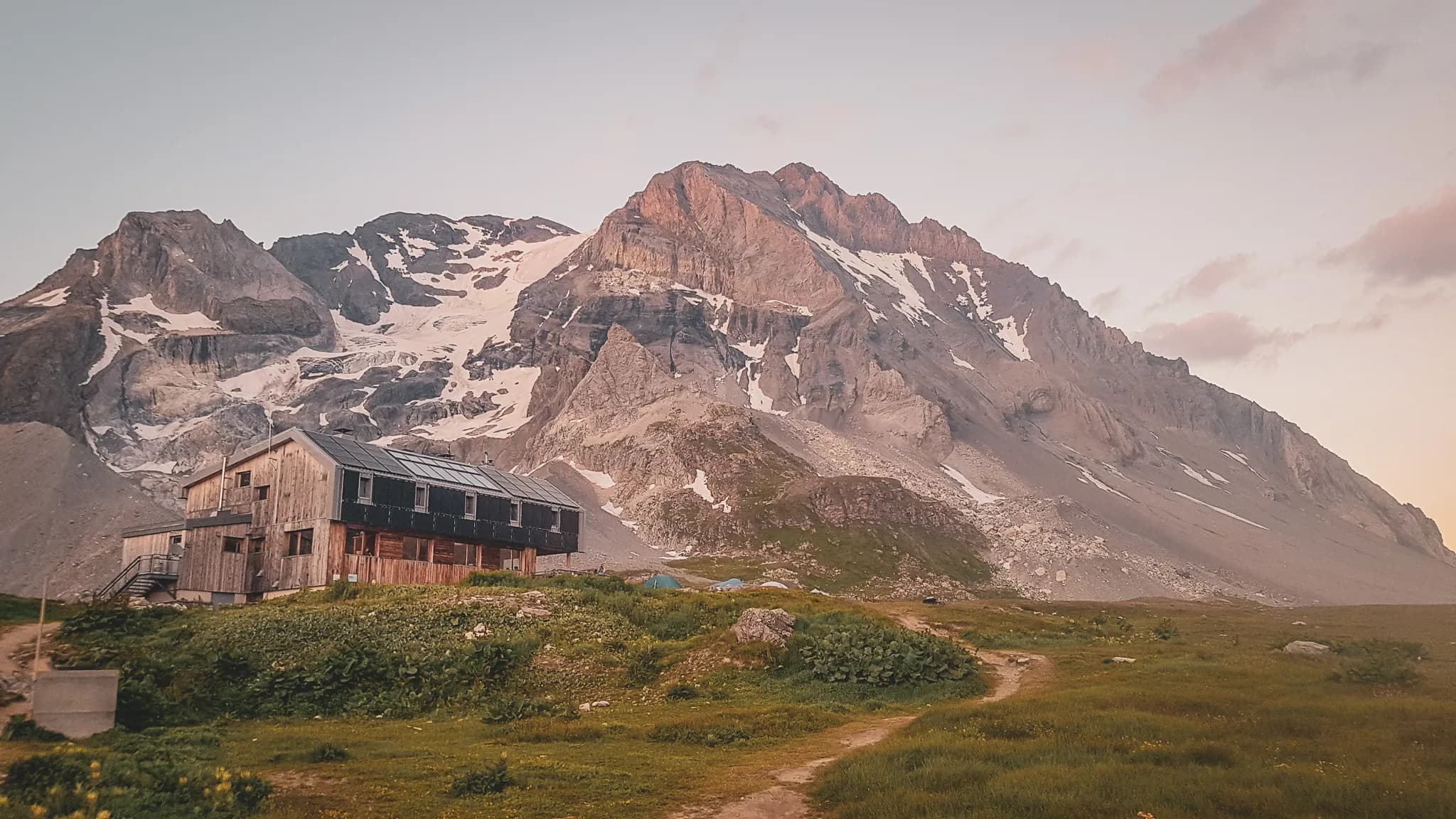 A wooden chalet sits at the foot of a majestic mountain, surrounded by natural scenery. The mountain, with its craggy peaks and areas of residual ice, stands proudly in the background. The sky