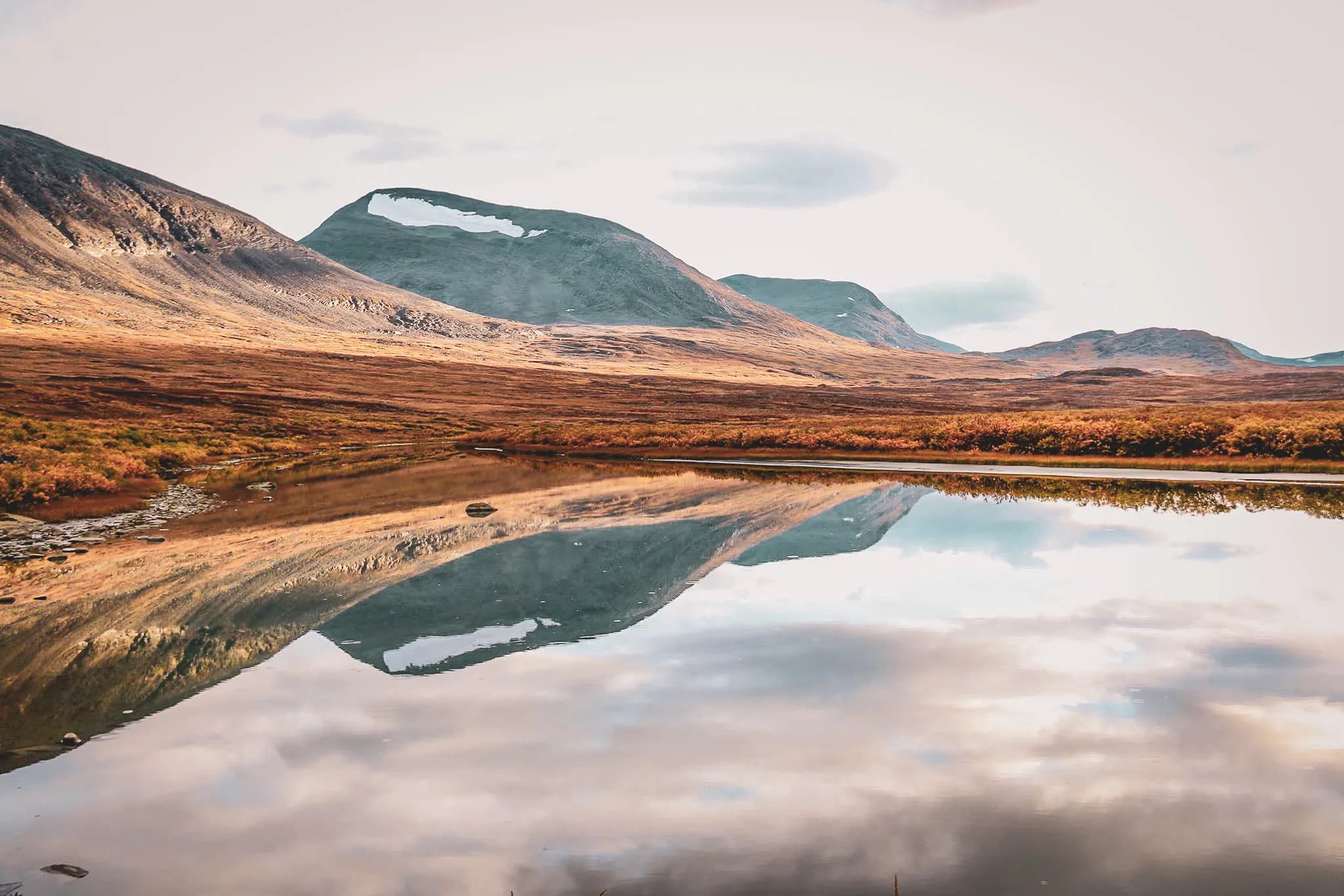 Un paysage naturel serein reflétant des montagnes majestueuses dans un étang calme. Les collines sont bordées de végétation automnale aux teintes dorées et orangées, tandis que les sommets portent encore quelques traces de neige.