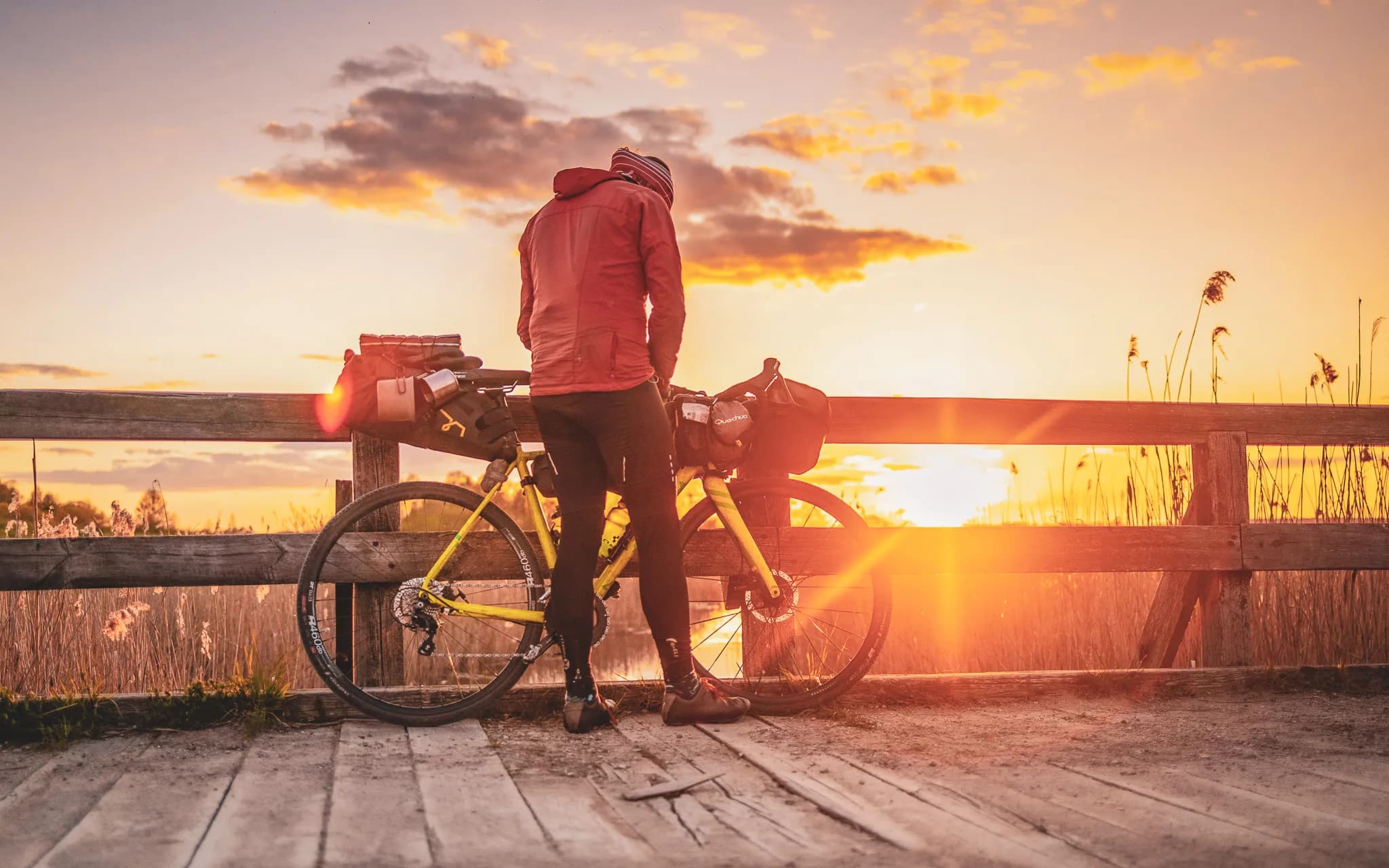 A cyclist in riding gear stands beside his yellow bike against a wooden fence, admiring a magnificent sunset. The surrounding landscape, with its tall grasses, is bathed in golden light.